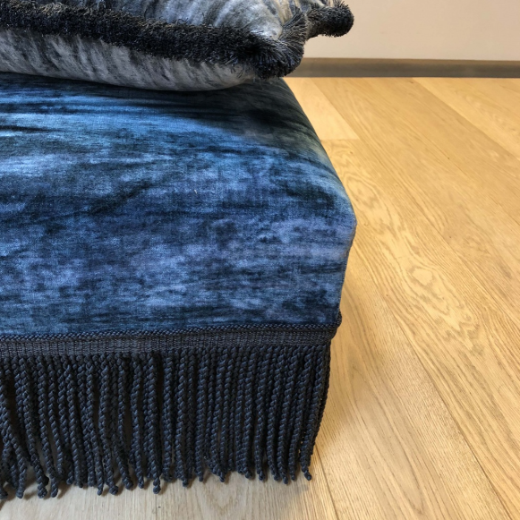 Blue textured ottoman with midnight baya bullion fringe and a grey pillow on a wooden floor.