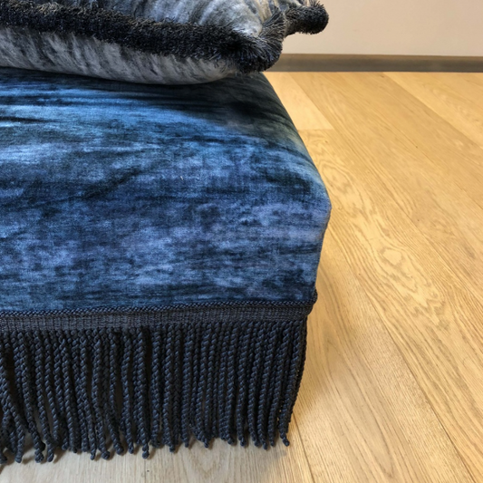 Blue textured ottoman with midnight baya bullion fringe and a grey pillow on a wooden floor.
